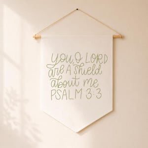 Op de afbeelding: Een crèmekleurige stoffen wandbanner met de saliegroene tekst: YOU, O LORD ARE A SHIELD ABOUT ME PSALM 3:3. De banner hangt aan een lichtgekleurde houten stok met een dun, natuurkleurig touwtje eraan bevestigd om de banner op te hangen.