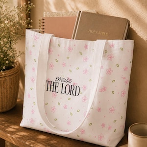 May include: A white canvas tote bag featuring a repeating pattern of small pink flowers and green leaves. The text "praise THE LORD" is printed in black on the front. The bag has two white fabric handles and contains a Holy Bible and a notebook.