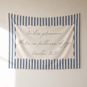 May include: A rectangular wall tapestry featuring a cream-coloured centre panel with a scalloped border, printed with the black cursive text: In his presence there is fullness of joy. Psalm 16:11. The background has vertical blue and white stripes.