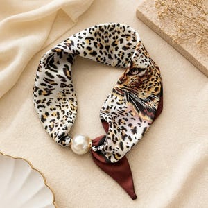 Puede incluir: Un pañuelo de cuello de aspecto sedoso con estampado de leopardo y un gráfico detallado de una cara de leopardo en un lado. El pañuelo se recoge a través de una cuenta grande y redonda de perla sintética color crema, con extremos marrón oscuro.