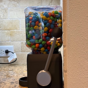 May include: A candy dispenser with a clear plastic container filled with multi-colored chocolate candies. The base is matte black with a silver circular dial and a long gray lever featuring a black spherical handle for dispensing the treats.