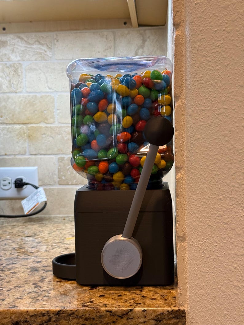 May include: A candy dispenser with a clear plastic container filled with multi-colored chocolate candies. The base is matte black with a silver circular dial and a long gray lever featuring a black spherical handle for dispensing the treats.