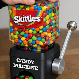 May include: A black candy machine with a clear plastic container filled with colorful Skittles. The machine features a silver metal dispensing lever on the side and a label on the front that reads CANDY MACHINE in white capital letters.