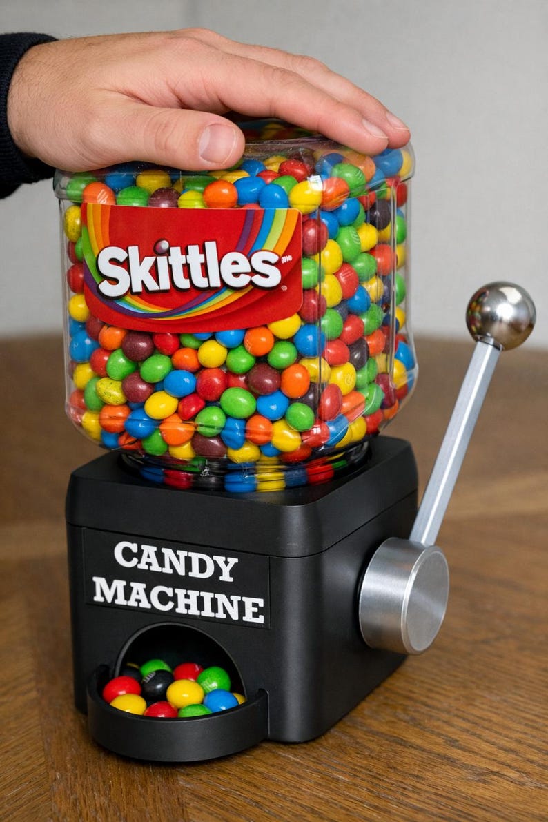 May include: A black candy machine with a clear plastic container filled with colorful Skittles. The machine features a silver metal dispensing lever on the side and a label on the front that reads CANDY MACHINE in white capital letters.