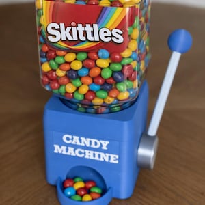 May include: A blue plastic candy machine with a clear square container filled with multi-colored Skittles. The front of the blue base features the white text CANDY MACHINE. A blue-tipped lever is attached to the side, and a small tray holds candies.