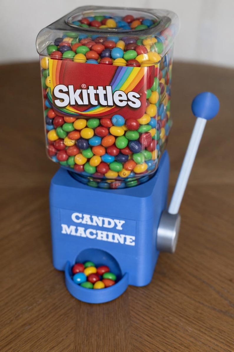 May include: A blue plastic candy machine with a clear square container filled with multi-colored Skittles. The front of the blue base features the white text CANDY MACHINE. A blue-tipped lever is attached to the side, and a small tray holds candies.