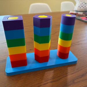 May include: A toy set featuring three vertical stacks of colorful blocks on a light blue rectangular base. The stacks consist of square, hexagonal, and cylindrical shapes, each featuring a rainbow sequence of purple, blue, green, yellow, orange, and red.