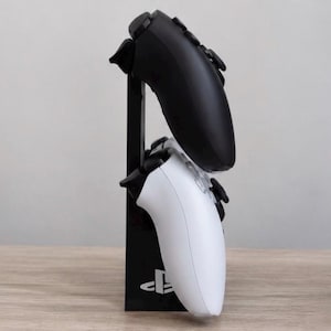 May include: A black vertical gaming controller stand featuring the PlayStation logo at the base. The stand holds one black wireless controller in the top position and one white wireless controller in the bottom position, both oriented vertically.