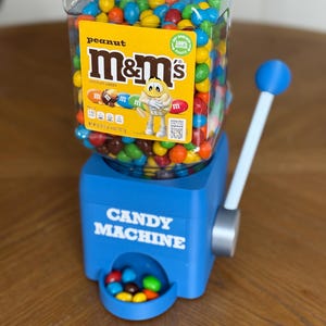 May include: A blue plastic candy machine with a clear square hopper filled with multi-colored peanut M&M's. The hopper features a yellow label with the M&M's logo and a character. The machine has a white lever with a blue knob and a dispensing tray.