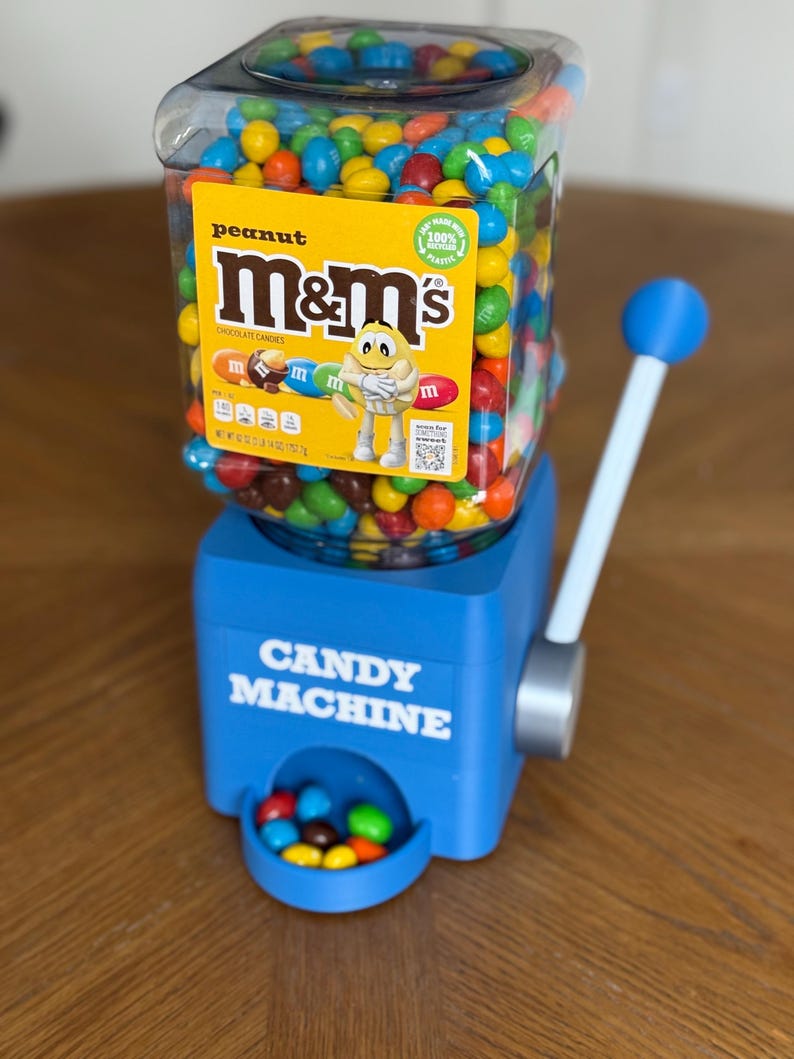 May include: A blue plastic candy machine with a clear square hopper filled with multi-colored peanut M&M's. The hopper features a yellow label with the M&M's logo and a character. The machine has a white lever with a blue knob and a dispensing tray.