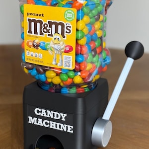 May include: A black candy machine with a clear plastic hopper filled with multi-colored peanut M&M's chocolate candies. The hopper features a yellow label with the M&M's logo and a yellow character. The base has the text CANDY MACHINE and a lever.