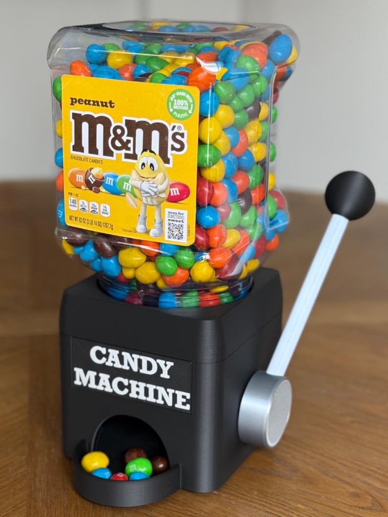 May include: A black candy machine with a clear plastic hopper filled with multi-colored peanut M&M's chocolate candies. The hopper features a yellow label with the M&M's logo and a yellow character. The base has the text CANDY MACHINE and a lever.