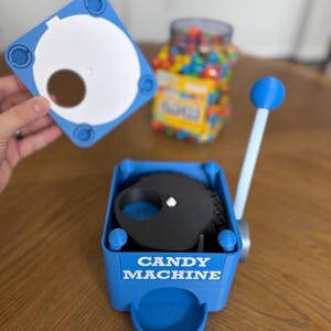 May include: A blue plastic candy machine with a white lever and a black internal gear mechanism. The front of the blue base features the white text CANDY MACHINE. A separate blue lid with a circular opening is shown above the main unit.