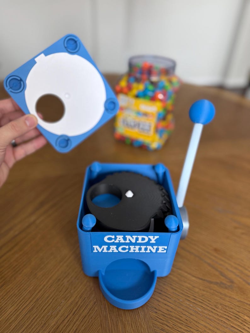 May include: A blue plastic candy machine with a white lever and a black internal gear mechanism. The front of the blue base features the white text CANDY MACHINE. A separate blue lid with a circular opening is shown above the main unit.
