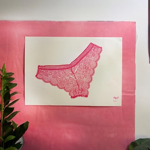 Peut inclure: Une impression d'art rectangulaire blanche présentant une illustration détaillée à l'encre rose vif de sous-vêtements en dentelle. L'impression affiche un motif à bords festonnés et inclut une signature manuscrite dans le coin inférieur.