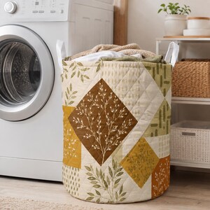 Könnte beinhalten: Ein zylindrischer, gesteppter Wäschekorb mit einem Patchwork-Design aus cremefarbenen, olivgrünen, senfgelben und rostorangen Quadraten. Der Stoff zeigt botanische Blatt- und Zweigmuster mit weißen Griffen an den Seiten.