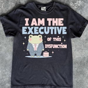 Może przedstawiać: Czarny t-shirt z krótkim rękawem z grafiką jasnozielonej żaby w szarym garniturze i różowym krawacie. Napis brzmi I AM THE EXECUTIVE OF THIS DYSFUNCTION jasnoniebieskimi i różowymi literami, ozdobiony małymi gwiazdkami i serduszkami.
