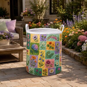Könnte beinhalten: Ein zylindrischer Wäschekorb aus gestepptem Stoff mit einem bunten Patchwork-Design aus verschiedenen botanischen Illustrationen wie Blumen, Blättern und Beeren. Der Korb hat ein weißes Innenfutter und zwei weiße Stoffgriffe an den Seiten.