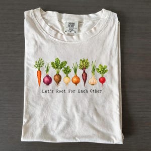 May include: A cream-coloured short-sleeve t-shirt featuring a graphic of various colourful root vegetables including a carrot, onions, and radishes. Below the vegetables, the text reads Let's Root For Each Other. The shirt has a Comfort Colors label.