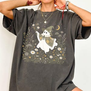 May include: A charcoal gray short-sleeve t-shirt featuring a graphic print of a white ghost wearing a green witch hat and a patterned cape. The ghost is surrounded by a field of white and yellow wildflowers, small stars, and a crescent moon.