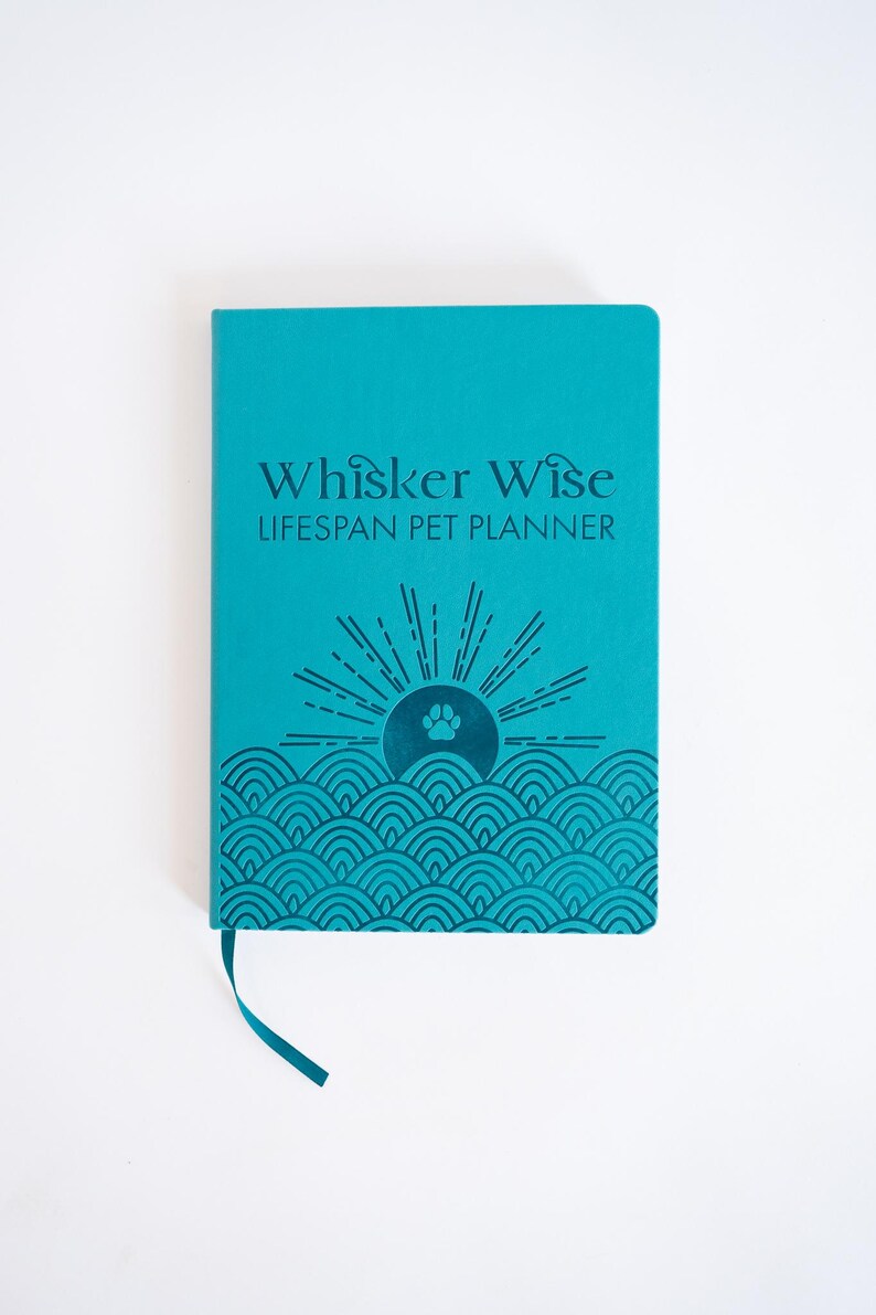 K&ouml;nnte beinhalten: Ein hellt&uuml;rkisfarbener Hardcover-Planer mit einem Lesezeichenband. Der Einband zeigt den Text "Whisker Wise LIFESPAN PET PLANNER" in Dunkelblau &uuml;ber einer Grafik einer aufgehenden Sonne &uuml;ber stilisierten Wellen mit einem Pfotenabdruck.