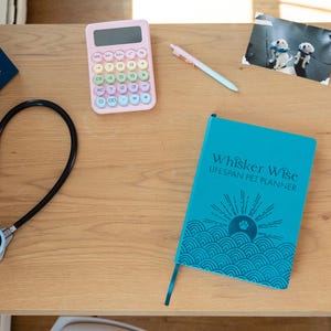 K&ouml;nnte beinhalten: Ein t&uuml;rkisfarbenes Haustier-Planer-Notizbuch mit dem Titel Whisker Wise Lifespan Pet Planner, verziert mit Sonne und Wellen, dazu ein schwarzes Stethoskop, ein pastellrosa Taschenrechner, ein hellblauer Stift, ein gerahmtes Foto und eine blaue Bulldoggenfigur.