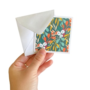May include: A white envelope with a green, orange, and white floral pattern card inside. The card has a dark green background with white flowers and orange leaves.
