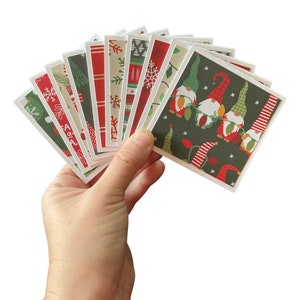 May include: A hand holding a fan of Christmas-themed square cards. The cards feature various festive patterns, including gnomes, snowflakes, and holiday lights, in red, green, and white. The cards are square with white borders.