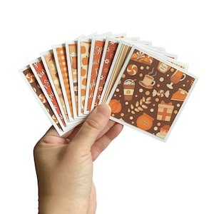 May include: A hand holds a fan of square cards with fall-themed designs. The cards feature patterns of pumpkins, coffee cups, and the words "Pumpkin Spice". Colors include orange, brown, and white, evoking autumn and seasonal themes.
