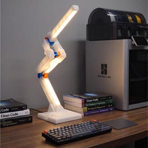 May include: A white articulated desk lamp with blue and orange accents is illuminated, casting a bright light. The lamp is on a wooden desk with a keyboard, books, and a 3D printer. The text "SOFTWARELIGHT.COM" is visible on the lamp base.