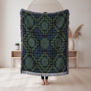 May include: A woven throw blanket featuring a repeating geometric pattern in shades of dark blue, forest green, and black. The design includes intricate spiral motifs centred within stepped square borders, finished with a fringed edge on all sides.