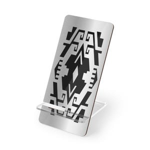 May include: A rectangular brushed metal phone stand featuring a bold black geometric pattern printed on the front surface. The stand is supported by a clear, transparent acrylic base with a small ledge designed to hold a mobile device securely.