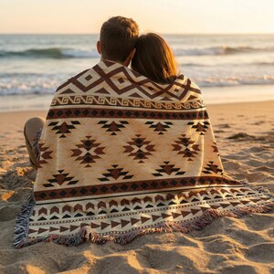 May include: A woven throw blanket featuring a repeating geometric pattern in shades of cream, brown, and black. The design includes diamond shapes, horizontal bands, and triangular motifs, finished with a fringed edge along the bottom border.