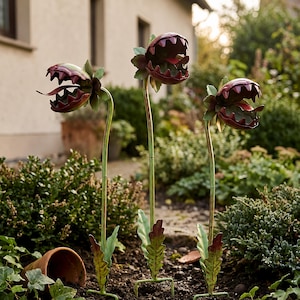 May include: Three metal garden stakes shaped like carnivorous plants with open, toothy mouths in deep red and green. Each plant features a long, slender green stem, jagged leaves at the base, and a wide, gaping head with a red tongue-like protrusion.