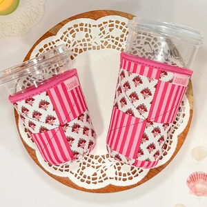 Puede incluir: Dos vasos de plástico transparente con tapa, cada uno envuelto en una funda de tela acolchada con un diseño de patchwork de rayas rosas y un estampado floral blanco con flores rojas. Cada funda tiene una etiqueta Saddler Shore.