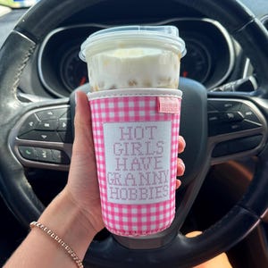 May include: A pink and white gingham fabric cup sleeve featuring a white rectangular patch with the text HOT GIRLS HAVE GRANNY HOBBIES in pink lettering. The sleeve is wrapped around a clear plastic cup filled with a creamy iced beverage.