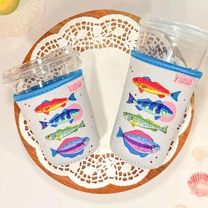 Puede incluir: Dos vasos de plástico transparente con tapa, cada uno dentro de una funda de neopreno gris claro con un patrón colorido de cuatro especies de peces. Cada funda tiene un borde azul y una pequeña etiqueta rosa con el texto Saddler Shore.
