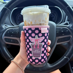 Puede incluir: Una funda de neopreno para taza en color azul marino con un patrón de lunares rosa brillante. La funda tiene un parche central que representa una taza de café con un lazo rosa y el texto girl breakfast. Una pequeña etiqueta dice Sadler Shore.