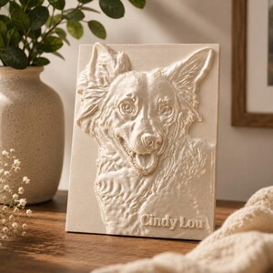 Puede incluir: Una escultura en relieve de color crema del rostro de un perro, con el nombre "Cindy Lou" en la parte inferior. El perro tiene una expresión feliz, con la boca abierta. La escultura está sobre una superficie de madera.