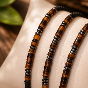 May include: A bracelet featuring alternating cylindrical tiger's eye beads and small metallic hematite-coloured spacers strung on a black braided cord. The design consists of repeating patterns of brown and black striped stone beads.