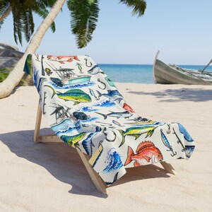 Peut inclure: Une serviette de plage blanche ornée d'un motif répétitif de poissons d'eau salée colorés, dont des coryphènes et des vivaneaux, ainsi que des illustrations de bateaux de pêche, de cannes, d'hameçons et de vagues bleues sur le tissu.