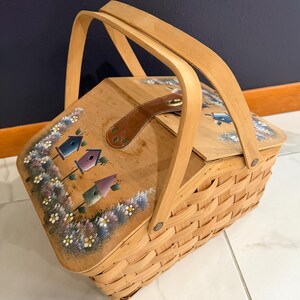 May include: A woven light wood basket with a hinged lid featuring a hand-painted design of three colorful birdhouses surrounded by small white flowers. The basket includes a brown leather strap closure and a sturdy arched wooden handle.