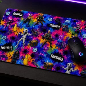 May include: A rectangular gaming mouse pad featuring a vibrant, multi-colored paint splatter background with various Fortnite characters, loot chests, and the Fortnite logo printed multiple times. A black wireless computer mouse rests on the surface.