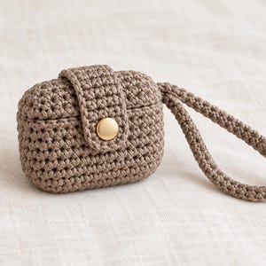 Könnte beinhalten: Ein taupefarbenes Häkeletui für kabellose Ohrhörer mit strukturiertem Strickmuster, einem sicheren Klappenverschluss mit einem glänzenden goldfarbenen Metalldruckknopf und einer passenden gehäkelten Handschlaufe zum bequemen Tragen.
