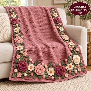 May include: A dusty rose crochet blanket featuring a textured stitch pattern with an elaborate border of three-dimensional crocheted flowers in shades of cream, light pink, and dark rose, accented with green leaves and small buds.
