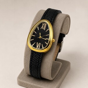 May include: A wristwatch featuring a teardrop-shaped gold-tone case and a black dial with gold-tone Roman numeral markers at the twelve and six o'clock positions. The watch has a textured black leather strap with a gold-tone buckle closure.