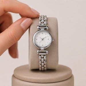 May include: A silver-toned analog wristwatch featuring a round white dial with small crystal hour markers and thin silver hands. The watch has a polished metal link bracelet with a textured, repeating oval pattern and a small side crown.