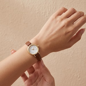 May include: A wristwatch featuring a round gold-tone case and a white dial with black Roman numeral hour markers. The watch is secured with a slim, textured brown leather strap that has visible stitching along the edges and a gold-tone buckle.