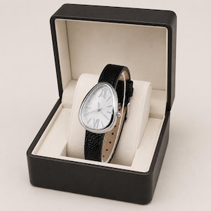 May include: A wristwatch with a teardrop-shaped silver case and a textured black leather strap, featuring a white dial with silver Roman numerals at the twelve and six o'clock positions, presented inside an open black hinged display box.