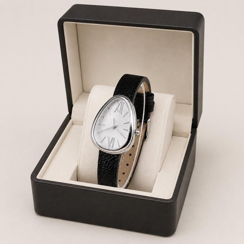 May include: A wristwatch with a teardrop-shaped silver case and a textured black leather strap, featuring a white dial with silver Roman numerals at the twelve and six o'clock positions, presented inside an open black hinged display box.